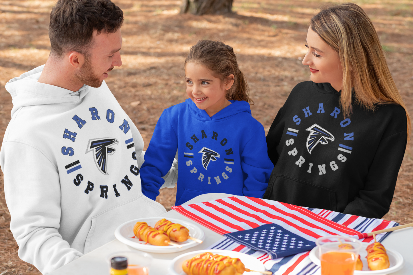 A father, mother and young daughter each wearing sponge fleece pullover hooded sweatshirts with Sharon Springs Falcons "Vintage Days" logo in black, white, and royal blue smiling an eating at a picnic table