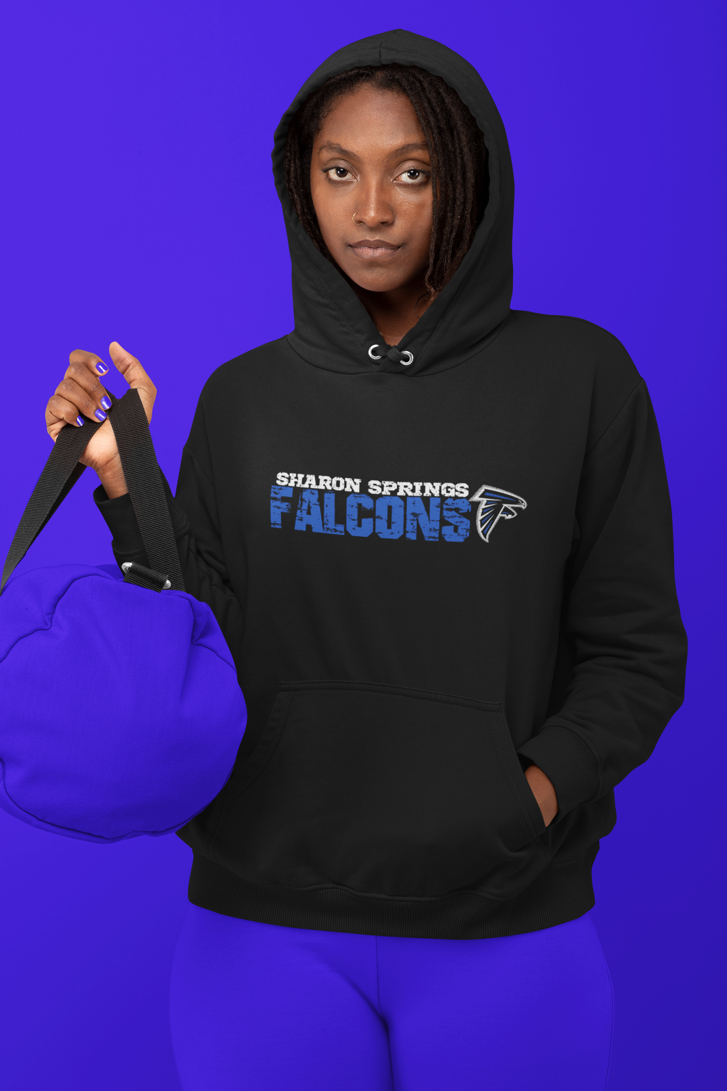 Woman wearing a black adult unisex sponge fleece pullover hooded sweatshirt with Sharon Springs Falcons “Distressed Logo” and blue pants and blue duffle bag standing against a solid blue surface