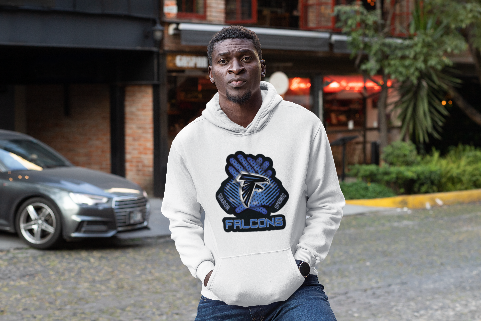 Man wearing a white adult unisex sponge fleece pullover hooded sweatshirt with Sharon Springs Falcons “Receiver's Gloves" logo standing in front of a building and car with hands in hoodie pockets