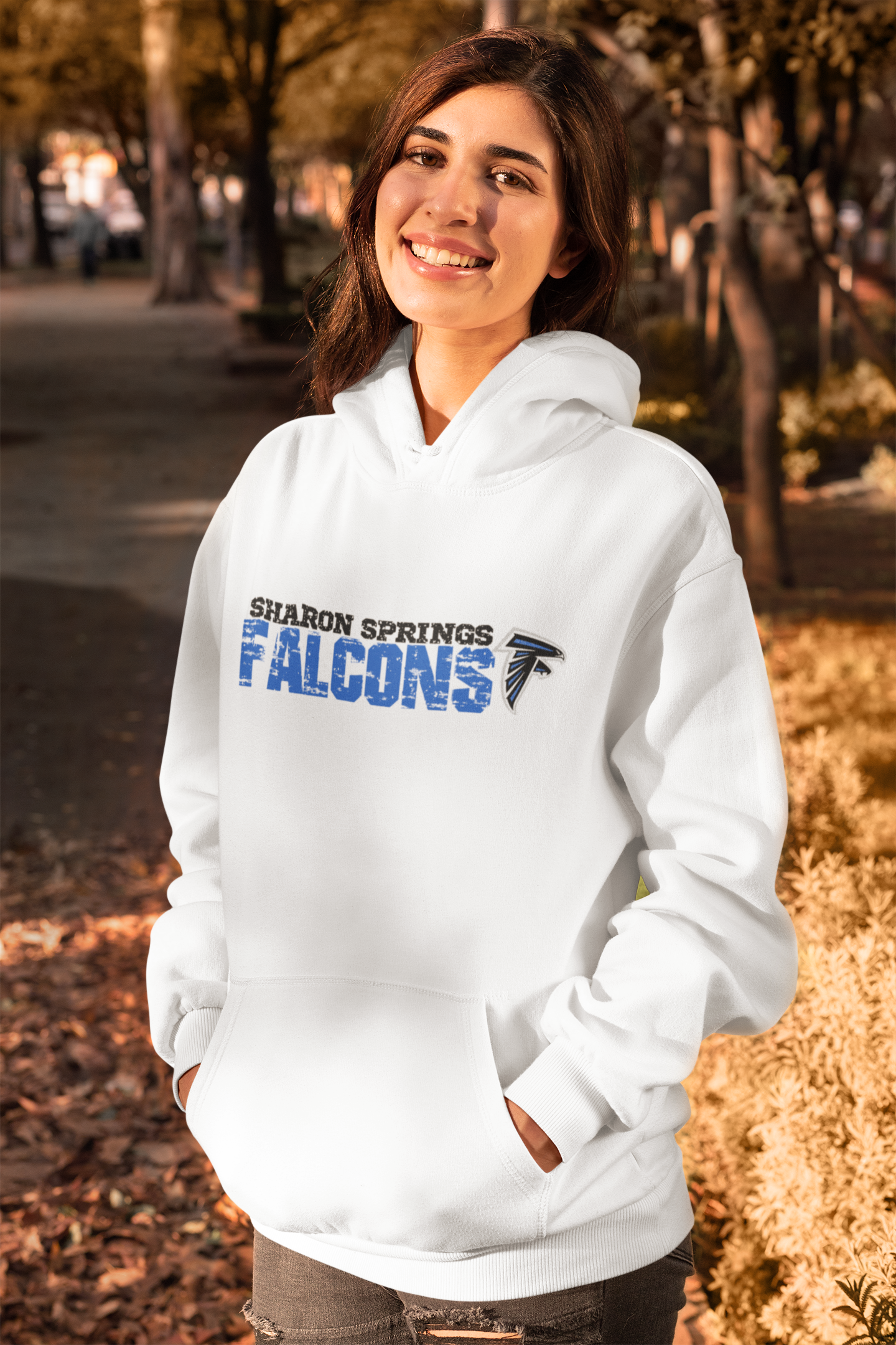 Smiling woman wearing a white adult unisex sponge fleece pullover hooded sweatshirt with Sharon Springs Falcons “Distressed Logo" and ripped jeans standing on the steet with her hands in her hoodie pockets in autumn. 