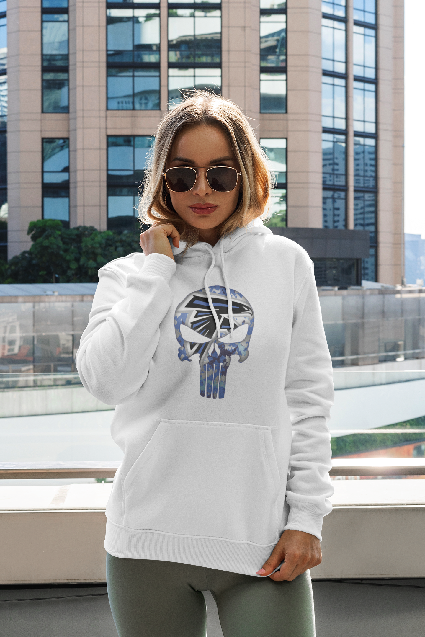 Woman wearing a white adult unisex sponge fleece pullover hooded sweatshirt with Sharon Springs Falcons “Punisher" logo, gray leggings and sunglasses standing in front of a building