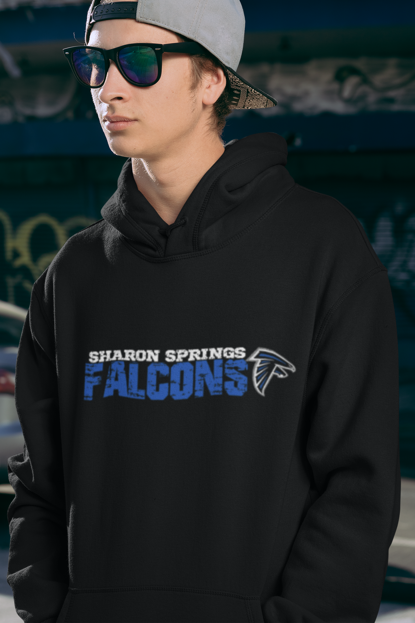 Young man wearing a black adult unisex sponge fleece pullover hooded sweatshirt with Sharon Springs Falcons “Distressed Logo", backwards had, and sunglasses standing with hands in hoodie pockets at skate park