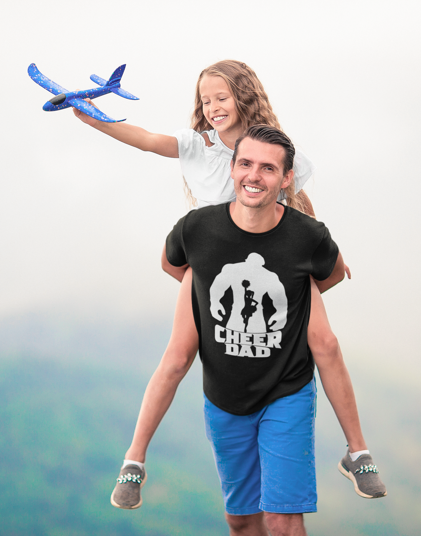 Man wearing black adult unisex t-shirt with Hulk "Cheer Dad" logo  and blue shorts giving his daughter a ride on his back while she holds a toy airplane